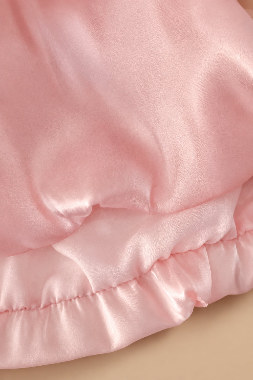 A close-up of light pink satin or silky fabric with soft folds and a ruffled hem, filling the frame.