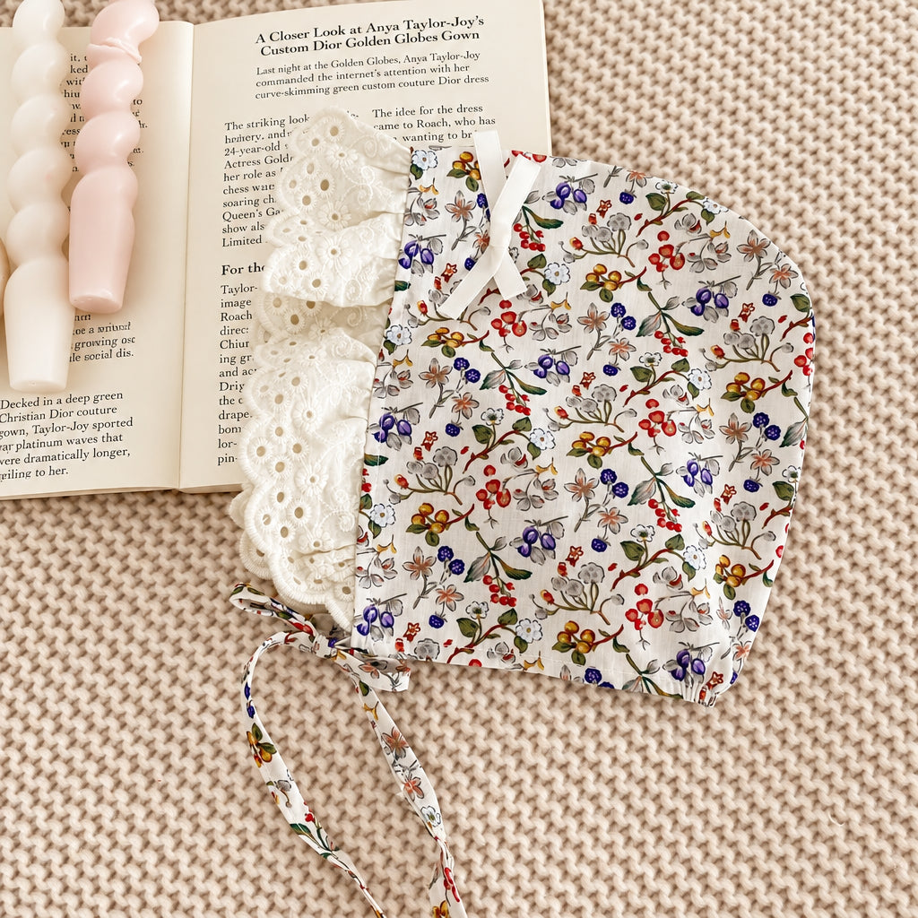 A close-up flat lay of a white fabric headband with a colorful floral and berry print, layered over a white eyelet lace headband, styled on a textured beige knit blanket with an open book and a pink beaded necklace in the background.