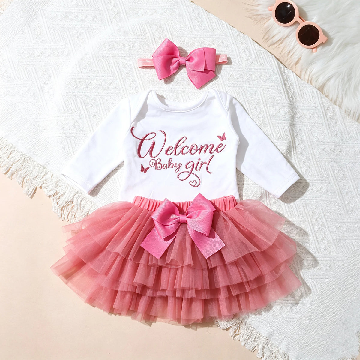 A baby girl's three-piece outfit set featuring a white long-sleeve top with 'Welcome Baby girl' printed in pink script, a pink tiered tulle tutu skirt with a large pink bow at the waist, and a matching pink bow headband. The set is styled in a flat lay on a white textured blanket with pink sunglasses in the corner.