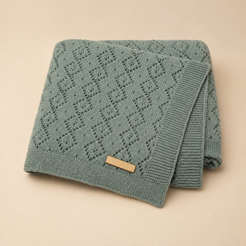 A folded sage green knit baby blanket with a textured diamond pattern and a small tan brand label, shown on a plain white background.