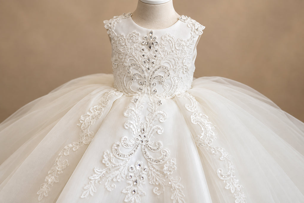 A close-up of a white or ivory formal dress displayed on a mannequin, featuring intricate lace embroidery, beaded embellishments, and a voluminous tulle skirt.