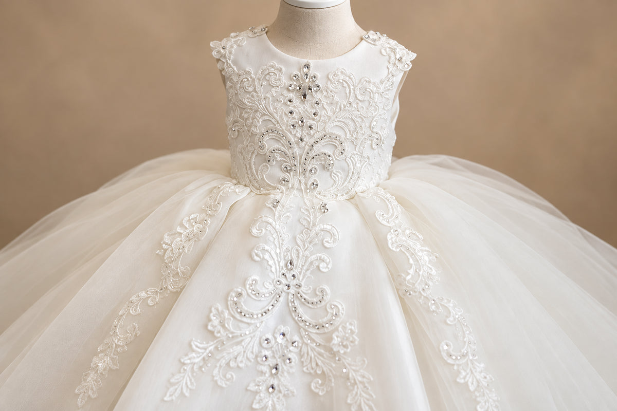 A close-up of a white or ivory formal dress displayed on a mannequin, featuring intricate lace embroidery, beaded embellishments, and a voluminous tulle skirt.