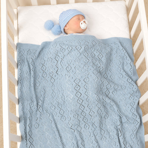A baby wearing a blue striped beanie with a pom-pom and a pacifier is lying in a white crib, covered with a light blue open-knit blanket.
