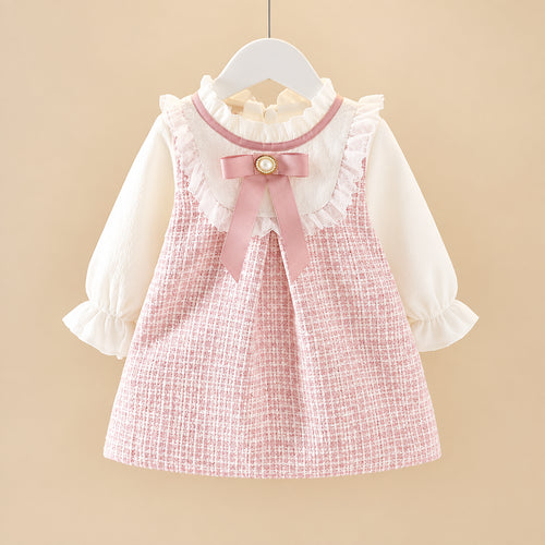 A toddler girl's two-piece outfit featuring a pink and white textured tweed pinafore vest with a pink bow and ruffled collar, layered over a white long-sleeve blouse with ruffled cuffs, hanging on a white hanger.