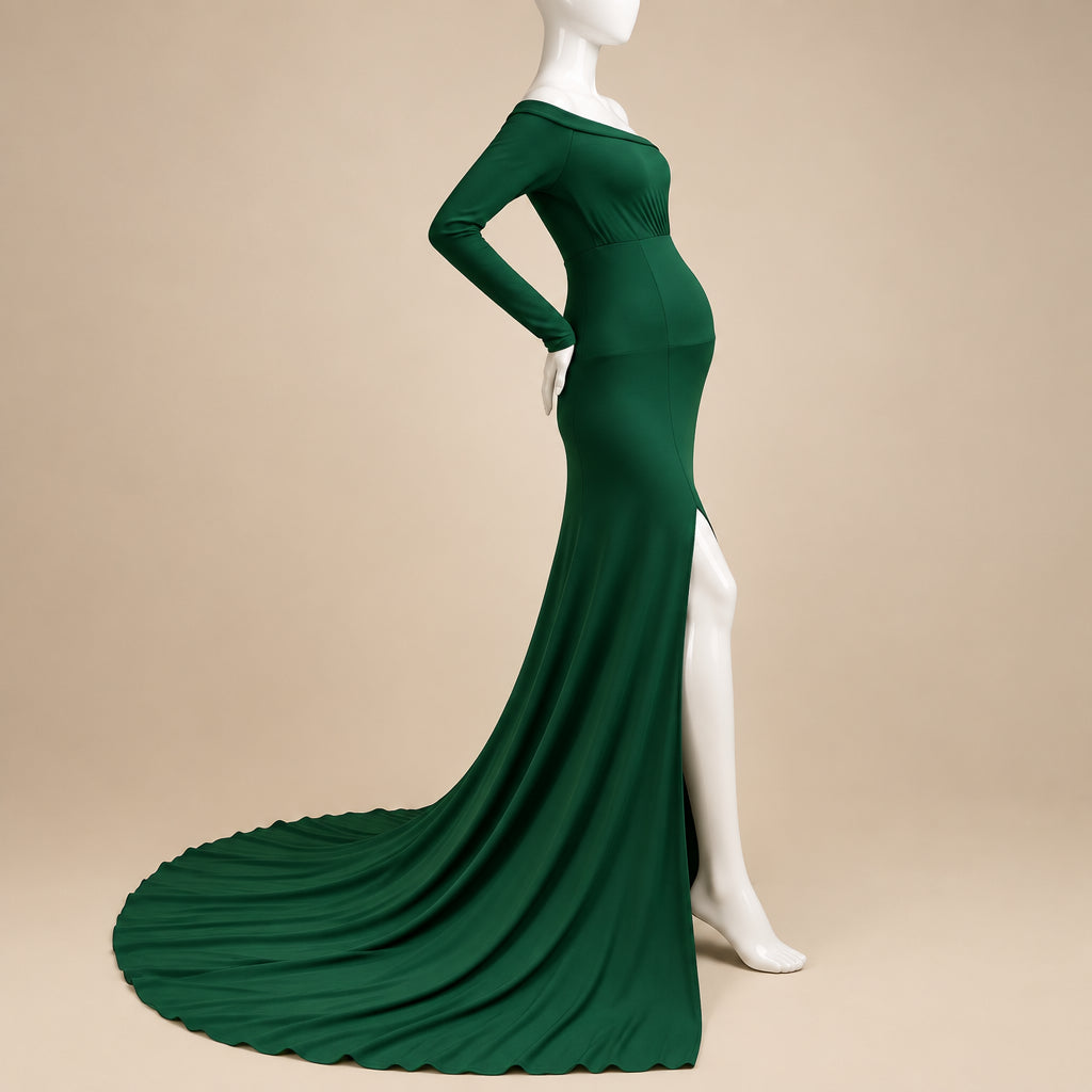 A long-sleeve, off-the-shoulder green maternity gown displayed on a white mannequin with a long train, set against a white wall and cream-colored rug.