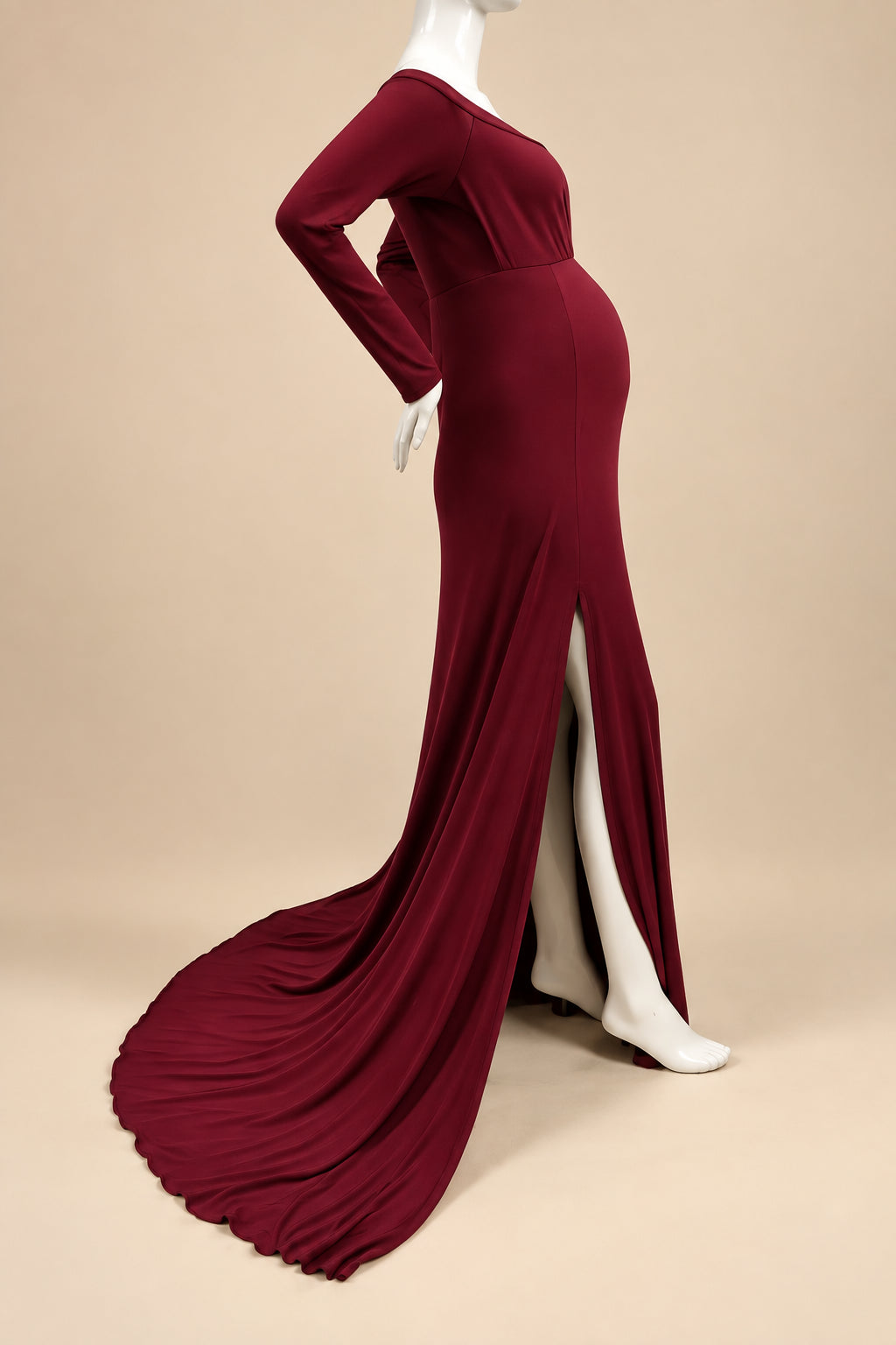 A long-sleeve, floor-length maternity gown in deep burgundy red, displayed on a white mannequin with a high leg slit, standing on a white shag rug against a white wall.