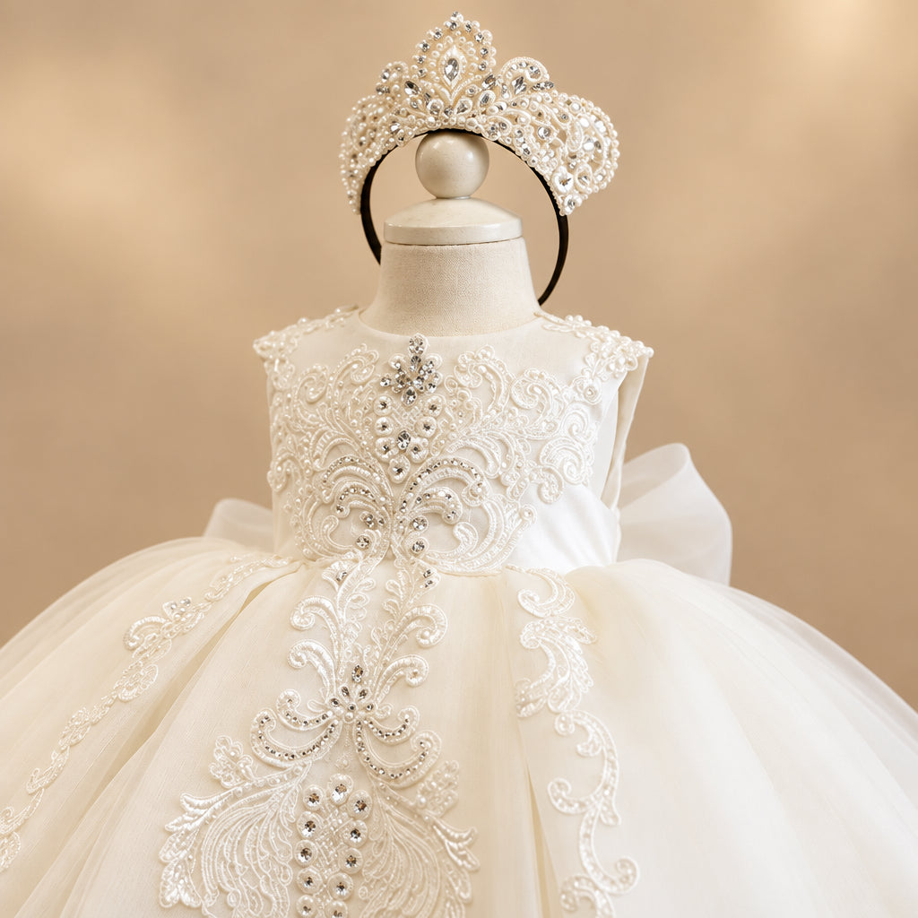 A white baby girl's formal dress and matching tiara set displayed on a white mannequin. The dress features intricate lace appliques, a ruffled skirt, and a matching jeweled tiara on a black headband.
