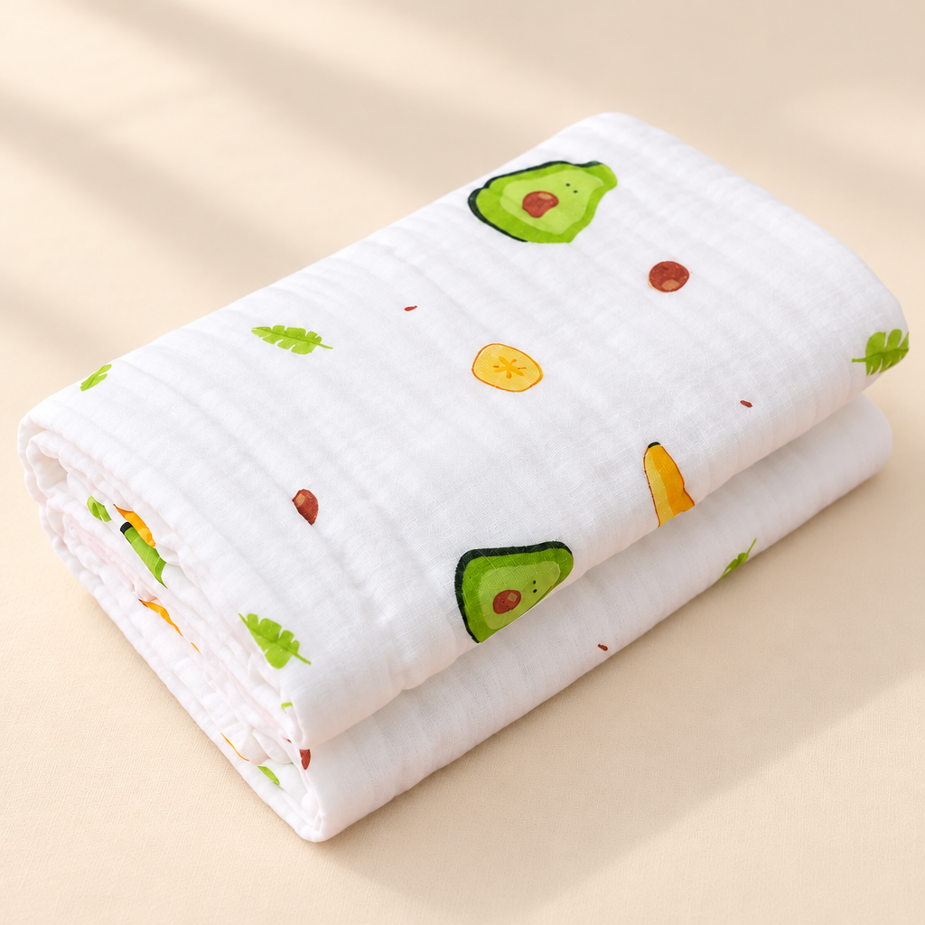 A folded white muslin baby blanket with a cute avocado and citrus print, displayed on a soft beige fabric background.