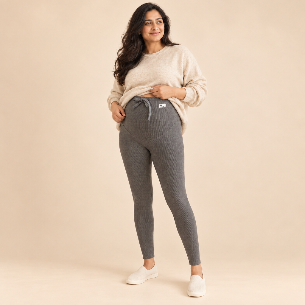 A woman modeling a pair of heather gray maternity leggings with a white drawstring and a small white logo patch, paired with a cream knit sweater and white slip-on shoes, against a plain off-white studio background.
